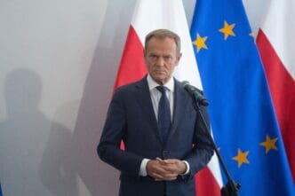 Donald Tusk, dressed in a navy suit, stands at a microphone with Polish and European Union flags visible behind him.