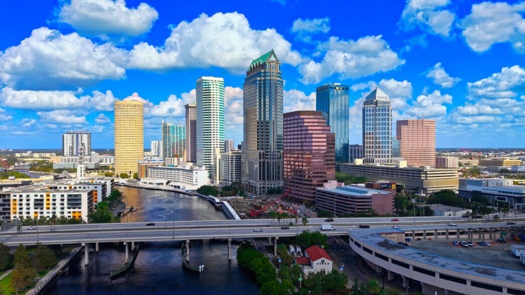 Aerial view of the downtown Tampa skyline featuring skyscrapers along the Hillsborough River under a bright, cloudy sky.