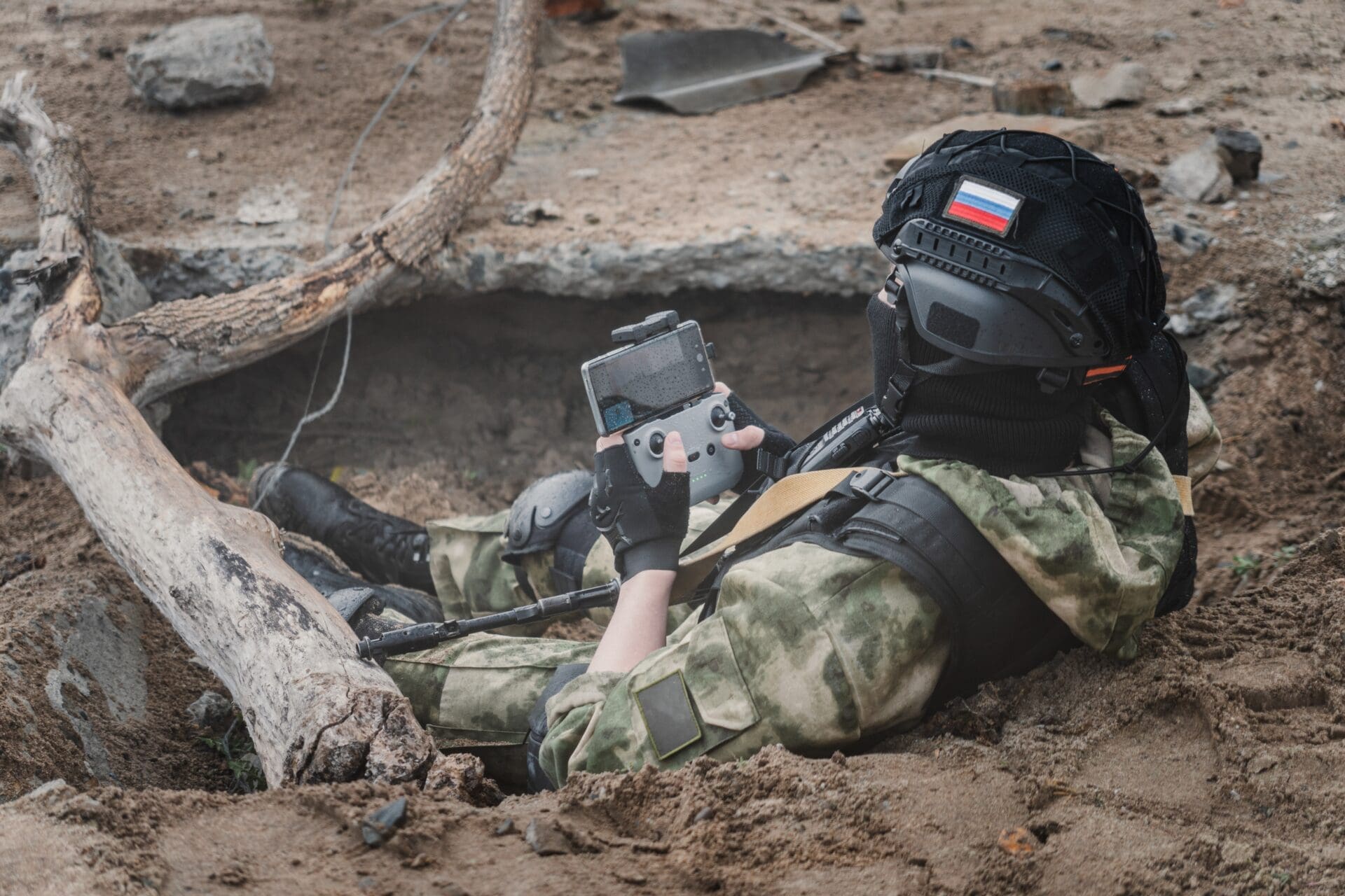 A soldier in camouflage lies in a trench, operating a drone remote control with a smartphone attached