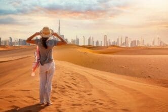 Woman in a straw hat stands on a sand dune, looking at the distant, modern Dubai skyline.