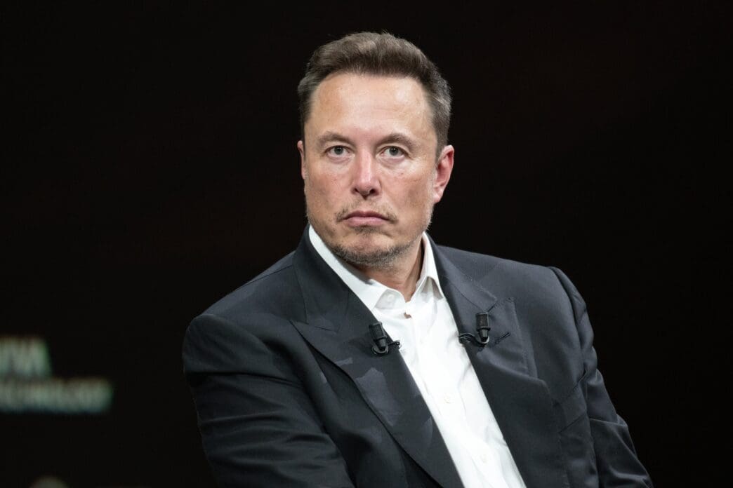 A close-up of Elon Musk in a suit, looking to the side against a dark background.