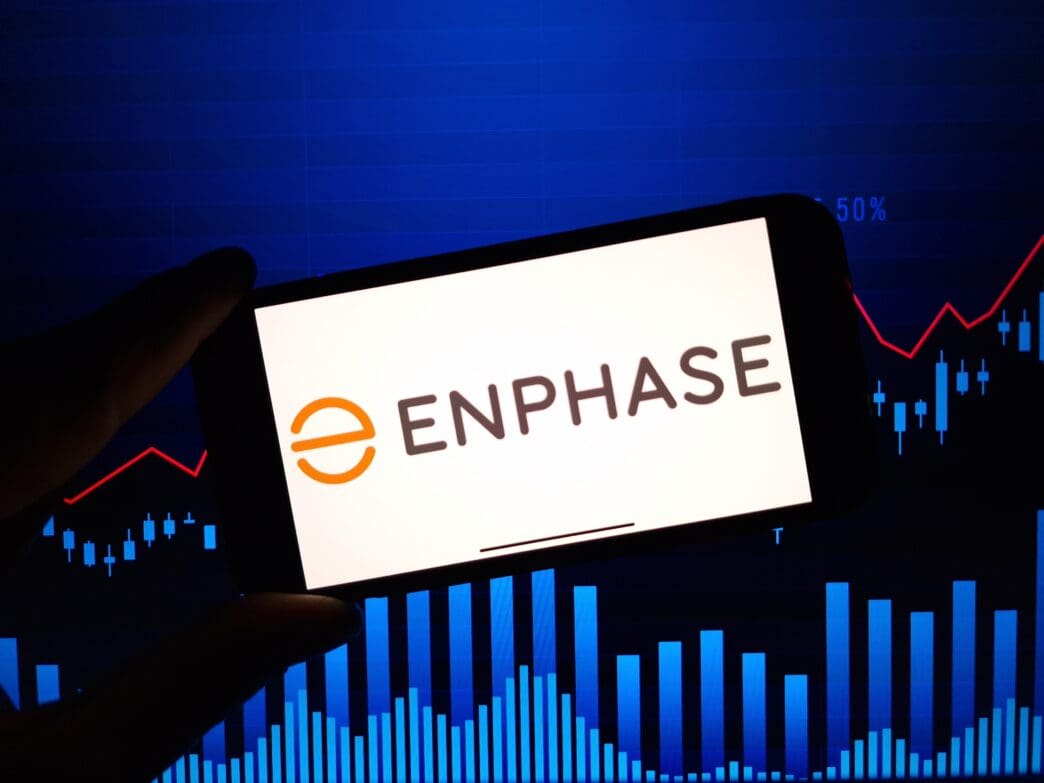A hand holds a smartphone displaying the Enphase Energy logo, with a red rising stock chart and blue bar graph visible in the background.