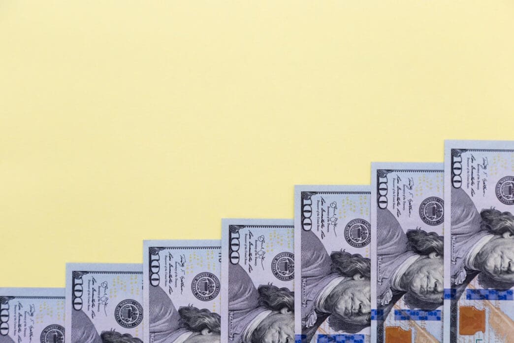 Seven US $100 bills are arranged in a rising staircase pattern on a solid light yellow background, suggesting growth.