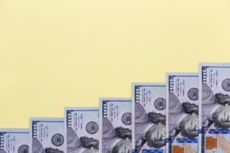 Seven US $100 bills are arranged in a rising staircase pattern on a solid light yellow background, suggesting growth.