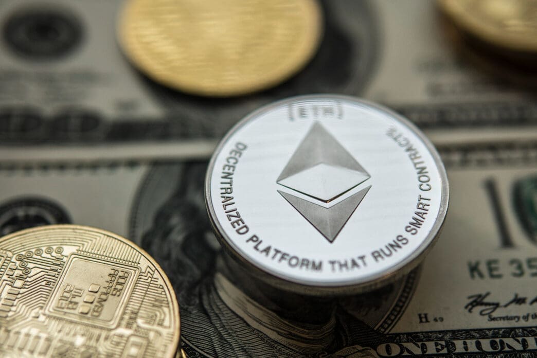 A silver Ethereum coin on top of a US one-hundred-dollar bill with other crypto coins nearby.