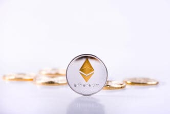 A physical silver coin with the golden Ethereum logo stands upright in sharp focus against a bright white background.