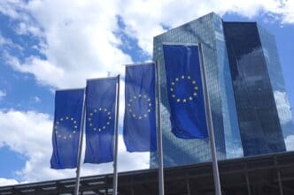Four European Union flags fly in front of the modern glass high-rise of the European Central Bank.