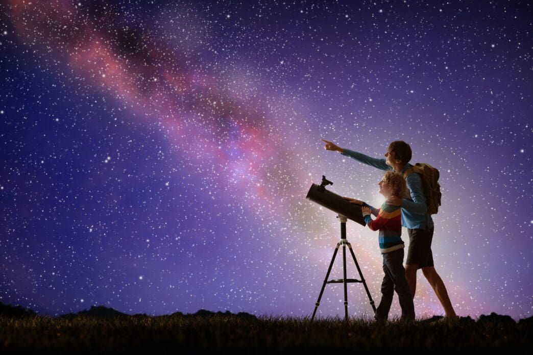 A man and a child look at the Milky Way through a telescope at night.