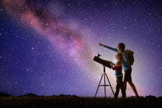 A man and a child look at the Milky Way through a telescope at night.