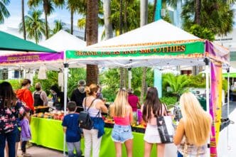 People shop at a colorful farmers market stall selling organic juices and fresh-cut fruits on a sunny street lined with palm trees.