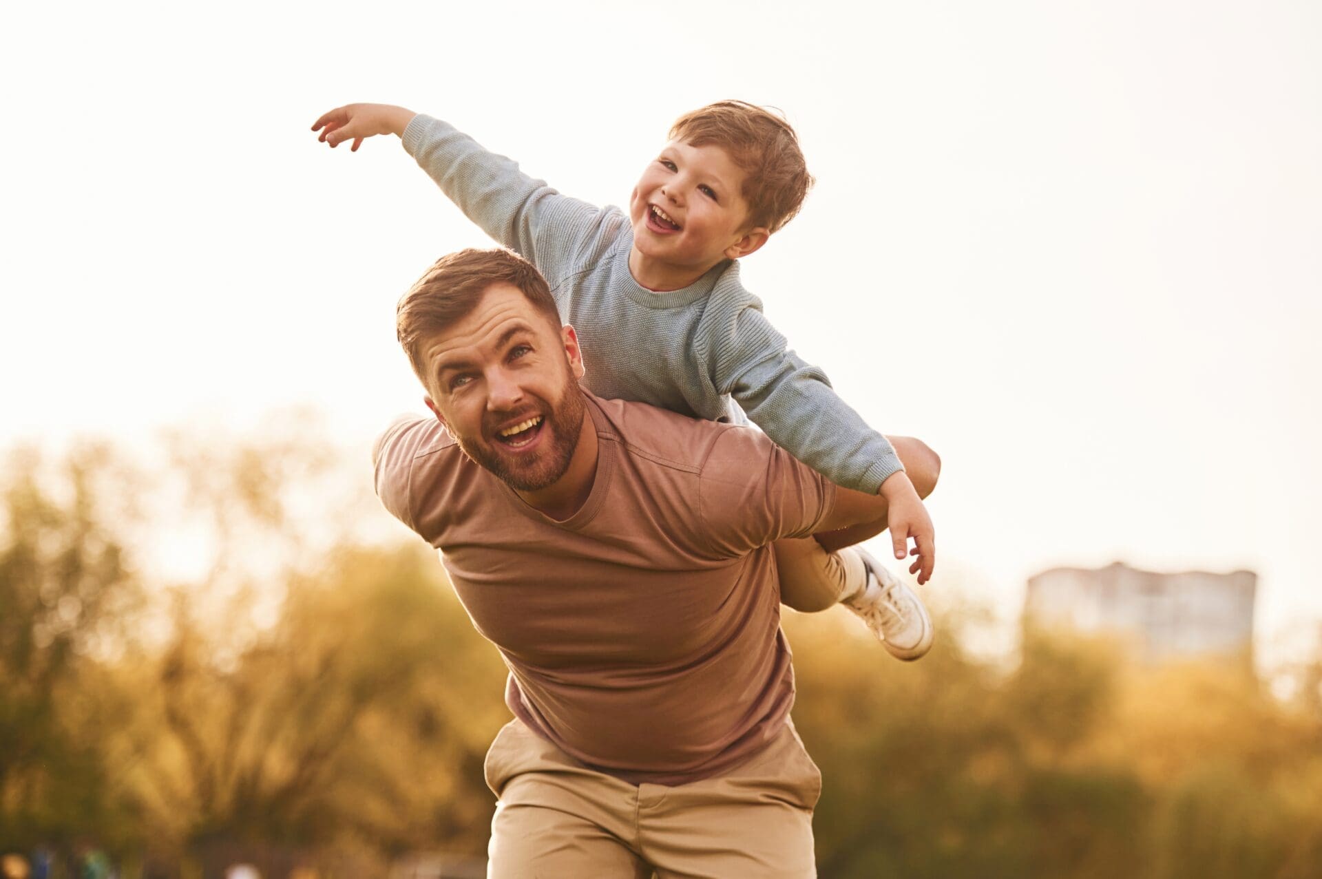 A father giving his joyful son a piggyback ride outdoors, with the boy spreading his arms to pretend he is flying