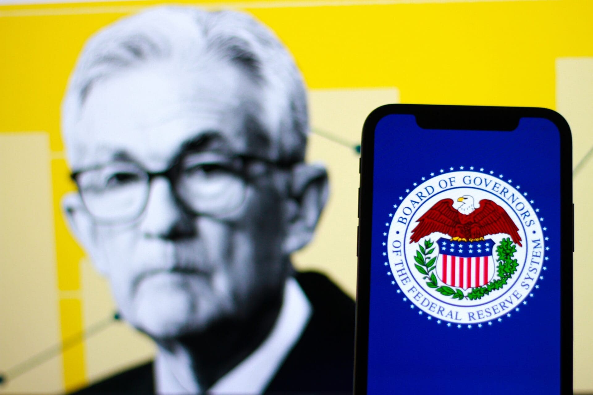 The Federal Reserve logo on a smartphone, with Fed Chair Jerome Powell in the background.