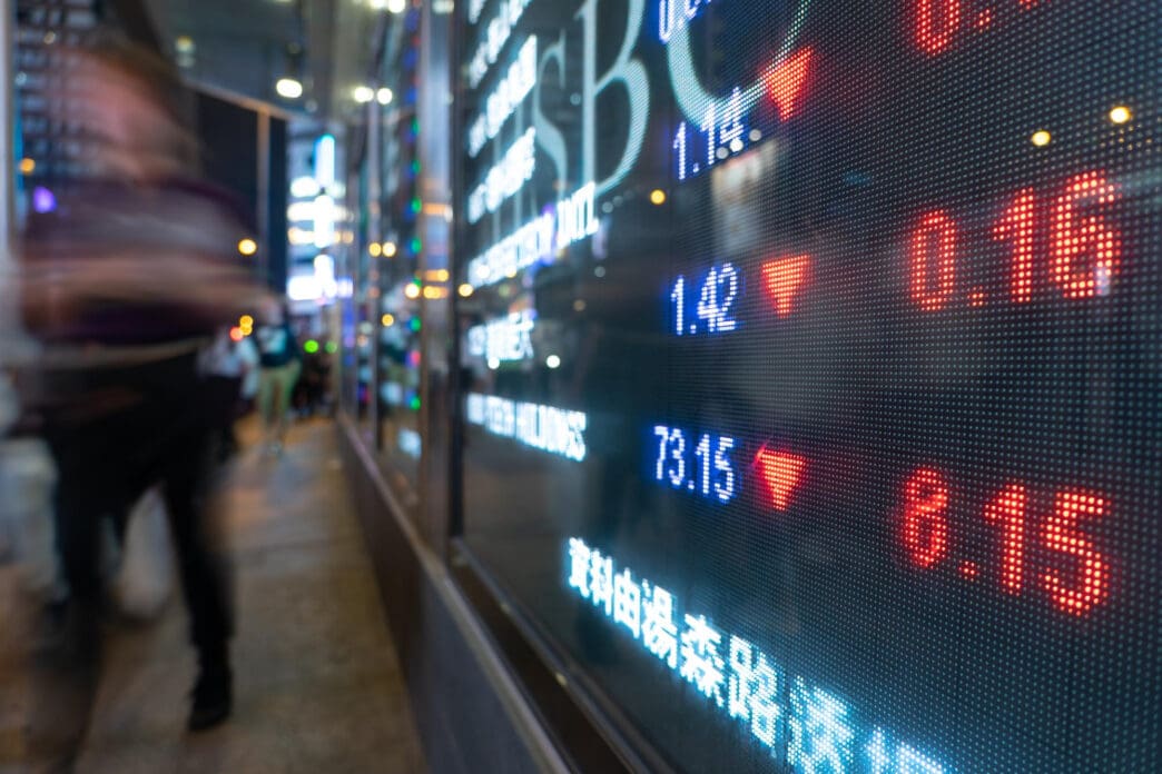 LED financial ticker screen displaying stock market data, showing mostly negative red numbers, viewed from a bustling city sidewalk at night.