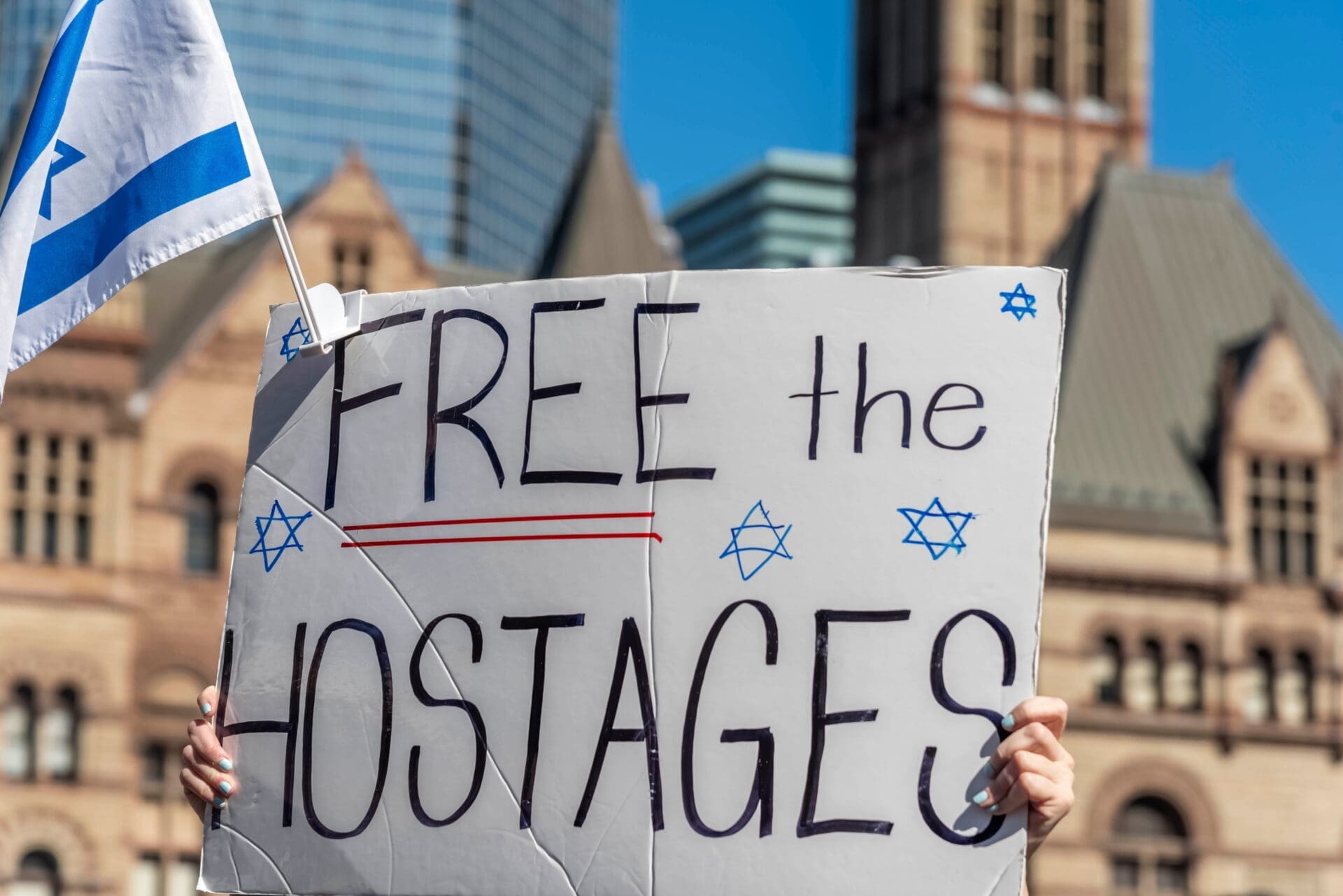 A person holding a sign that reads "FREE the HOSTAGES," with a small Israeli flag waving above it, at a pro-Israel rally