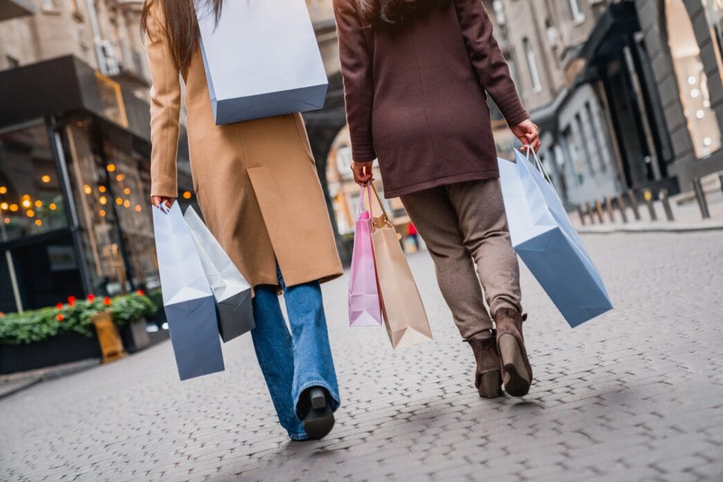 Two women in coats walking away on a city street, carrying multiple shopping bags in their hands.
