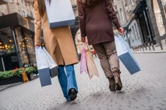 Two women in coats walking away on a city street, carrying multiple shopping bags in their hands.