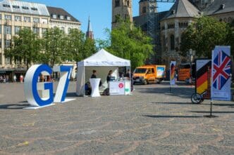 A large "G7" sign stands in a sunny European plaza with an information tent, an orange van, and a church in the background, promoting a finance meeting.