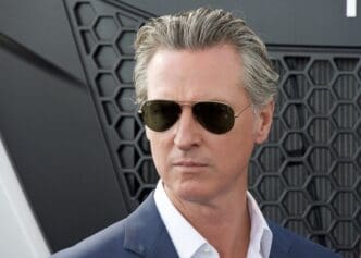 A close-up of Gavin Newsom wearing aviator sunglasses and a blue suit.