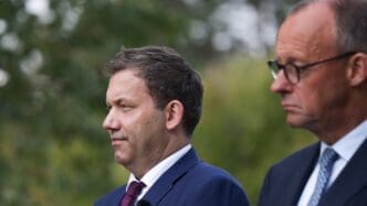 Close-up profiles of German Federal Finance Minister Lars Klingbeil (left) and Federal Chancellor Friedrich Merz (right) at an outdoor event.