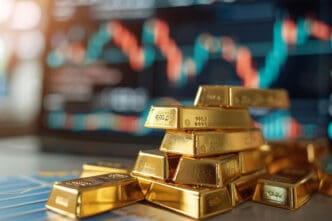 A stack of small, shiny gold bars rests in front of a blurred screen displaying a volatile financial chart.