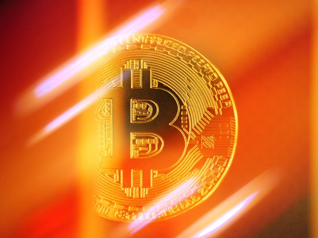 Close-up of a gold Bitcoin coin brightly lit by a golden-orange and red abstract background with glowing streaks.