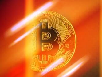 Close-up of a gold Bitcoin coin brightly lit by a golden-orange and red abstract background with glowing streaks.