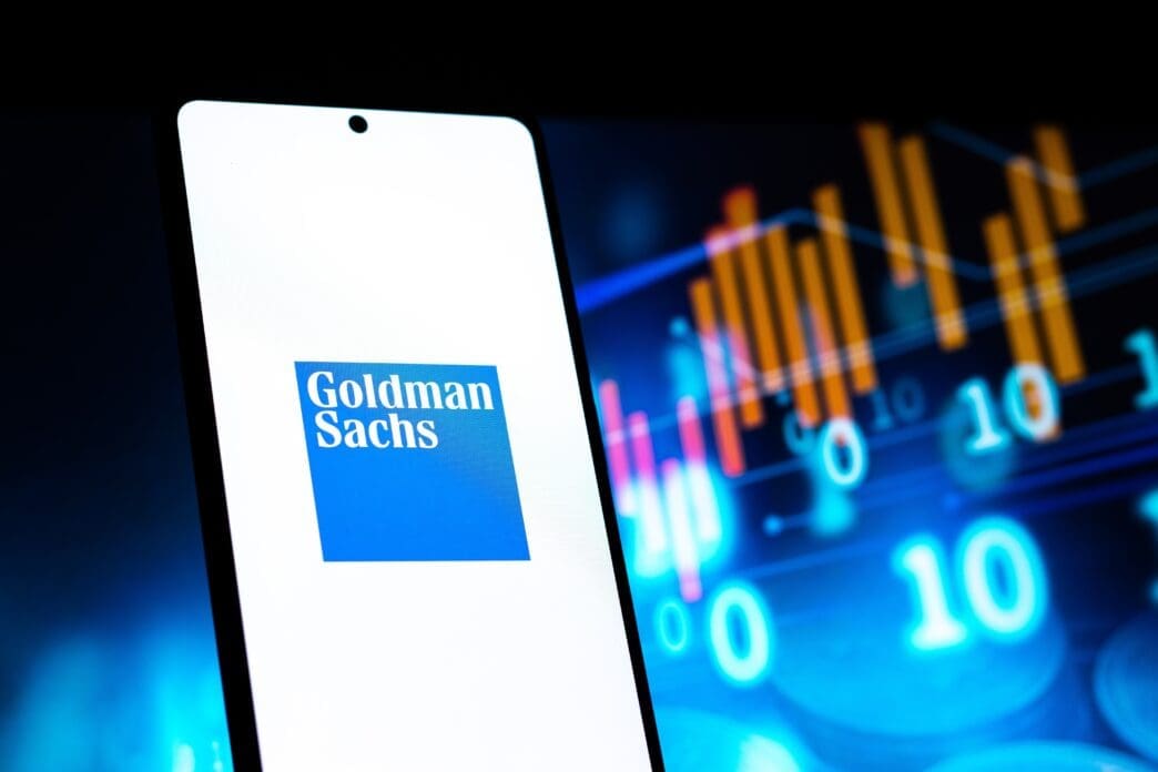 The Goldman Sachs logo is displayed on a smartphone screen, with blurred digital stock charts and numbers visible in the dark background.