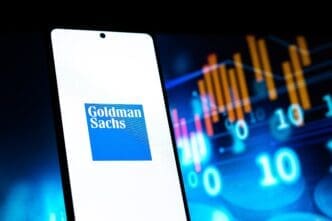 The Goldman Sachs logo is displayed on a smartphone screen, with blurred digital stock charts and numbers visible in the dark background.