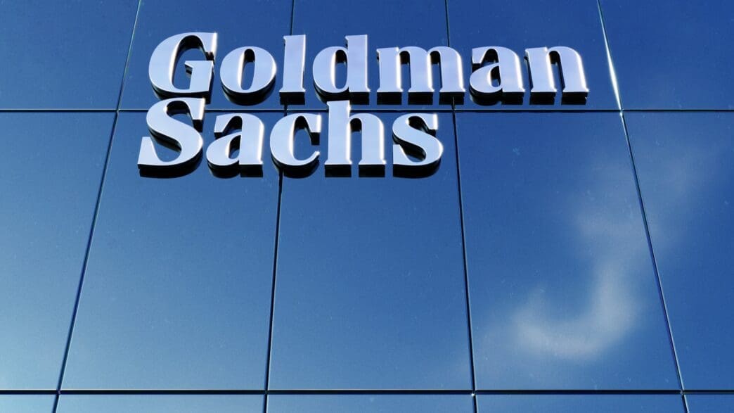 Goldman Sachs logo in silver letters mounted on a reflective blue glass facade.