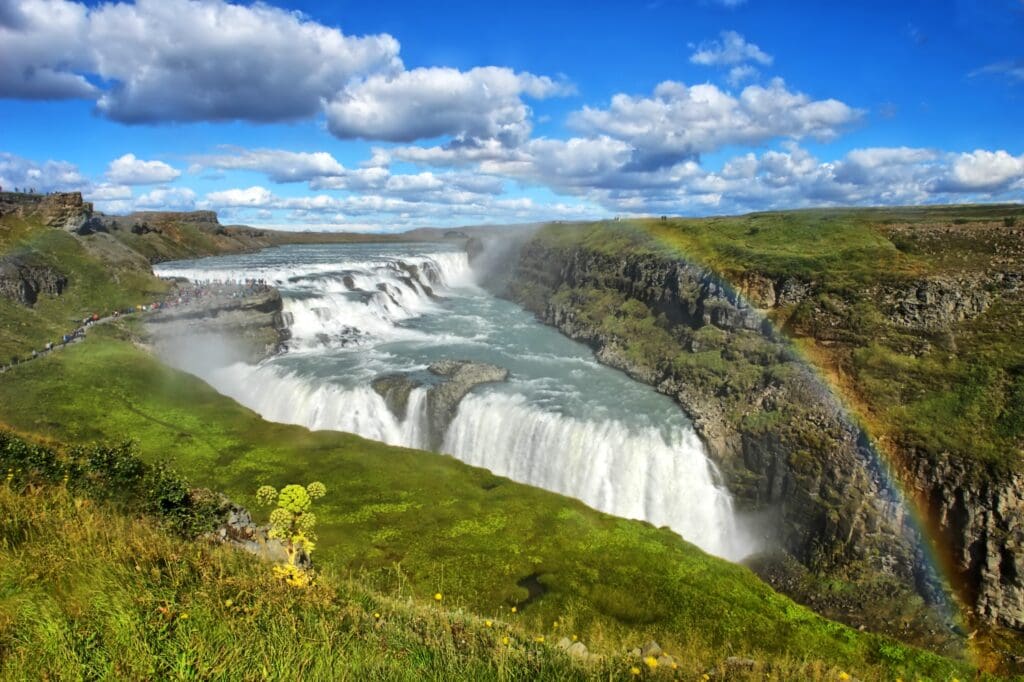 A stunning wide view of Gullfoss waterfall in Iceland, showing its two tiers, a crowd of tourists, and a bright rainbow in the mist.