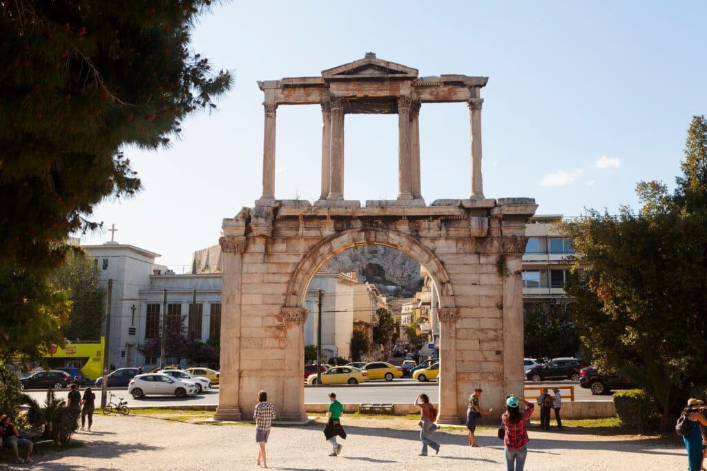The ancient Arch of Hadrian in Athens, Greece, with pedestrians nearby and city traffic passing behind it on a sunny day.