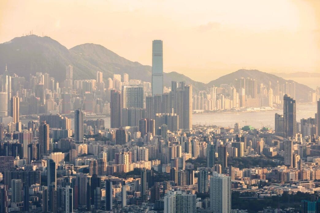 An aerial view of the dense Hong Kong skyline at sunset, with the iconic ICC tower rising above the city and hazy mountains.