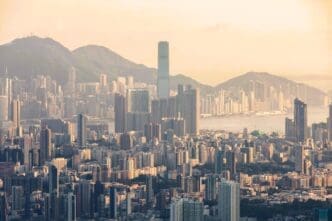 An aerial view of the dense Hong Kong skyline at sunset, with the iconic ICC tower rising above the city and hazy mountains.
