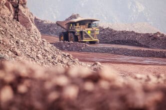 Massive haul truck with a raised bed driving on a dusty, reddish dirt road between piles of raw earth and rock at an open-pit mine.