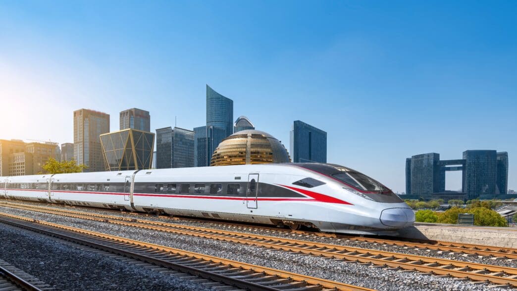 A high-speed train travels on tracks past a modern city skyline with futuristic buildings.