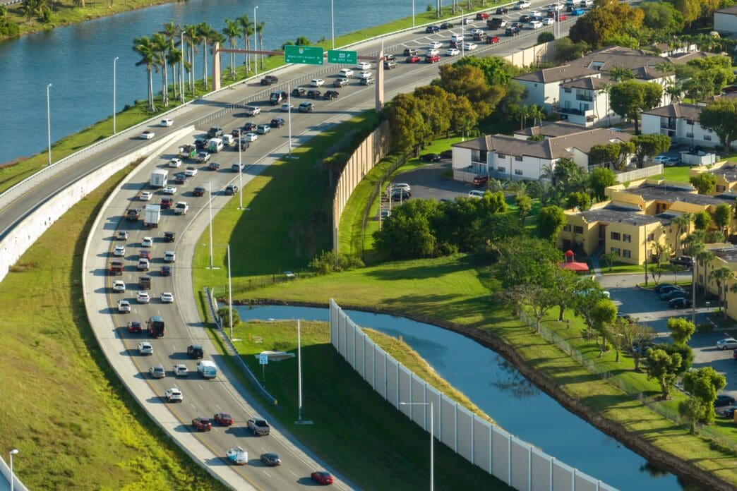 Aerial view of a busy curved highway next to a blue canal and residential apartment buildings in a sunny Miami suburb.