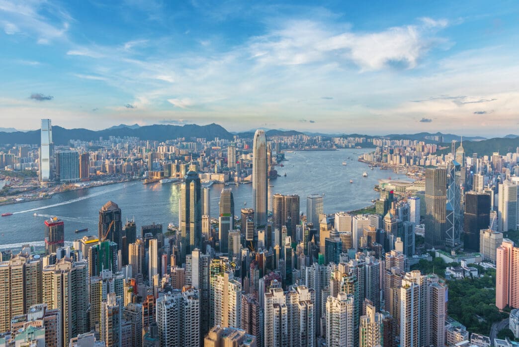 Panoramic view of Hong Kong's dense urban skyline, with towering skyscrapers surrounding Victoria Harbour under a blue sky.