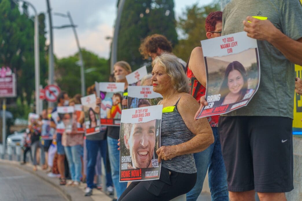 People stand on a sidewalk holding posters with photos of hostages and "Bring Him Home Now!".