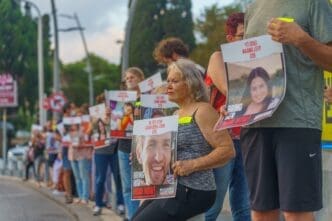 People stand on a sidewalk holding posters with photos of hostages and "Bring Him Home Now!".