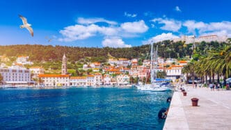 Hvar town harbor in Croatia, showing white buildings with orange roofs, sailboats, a seaside promenade, and a hilltop fortress under a bright blue sky with a flying seagull.