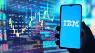 Hand holding a smartphone displaying the IBM logo against a background of digital stock charts.