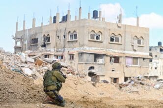 An IDF soldier kneeling behind a mound of dirt, aiming a rifle at a partially ruined concrete building in an area of destruction
