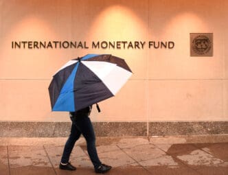 Pedestrian walking with a large black, blue, and white umbrella past the light-colored exterior wall of the International Monetary Fund building.