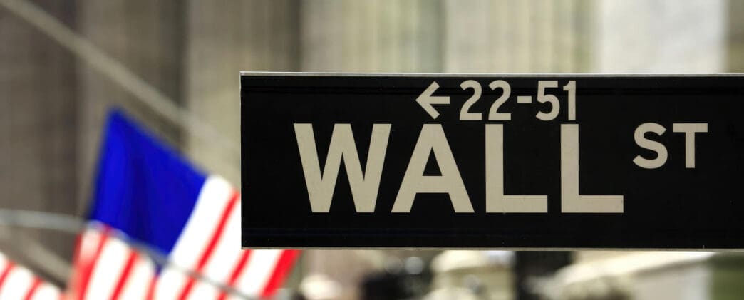 Close-up of the black and white Wall Street street sign, with an American flag blurred in the background.