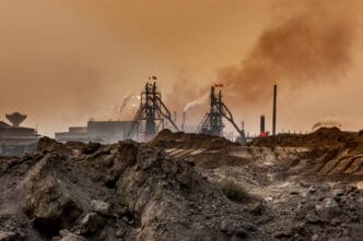 Dark industrial steel mill towers spewing thick smoke into a hazy, orange sky above piles of polluted earth.