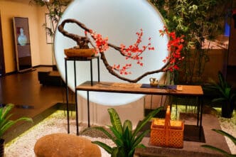 An indoor installation of a winding bonsai branch with small red leaves placed on a table with a teapot, all framed by a large circular light and white pebbles.