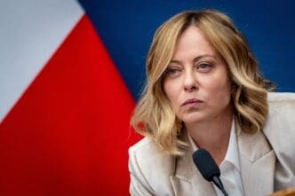 Close-up of Italian Premier Giorgia Meloni with a serious expression, speaking at a microphone with the Italian flag visible.