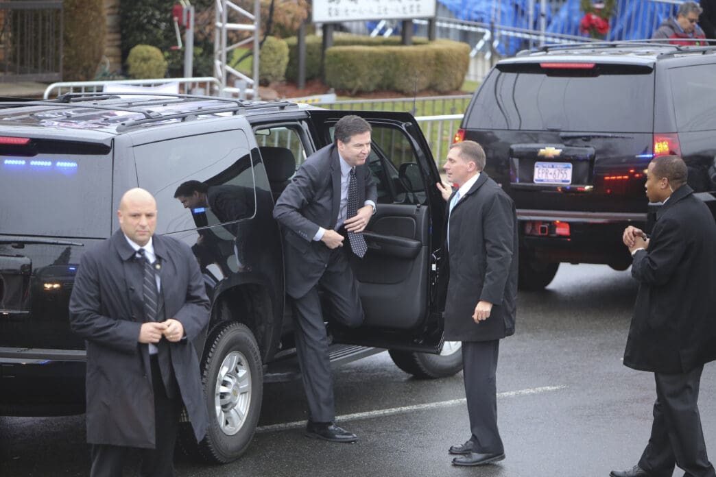 James Comey stepping out of a black SUV and speaking with another man, surrounded by security personnel and vehicles.