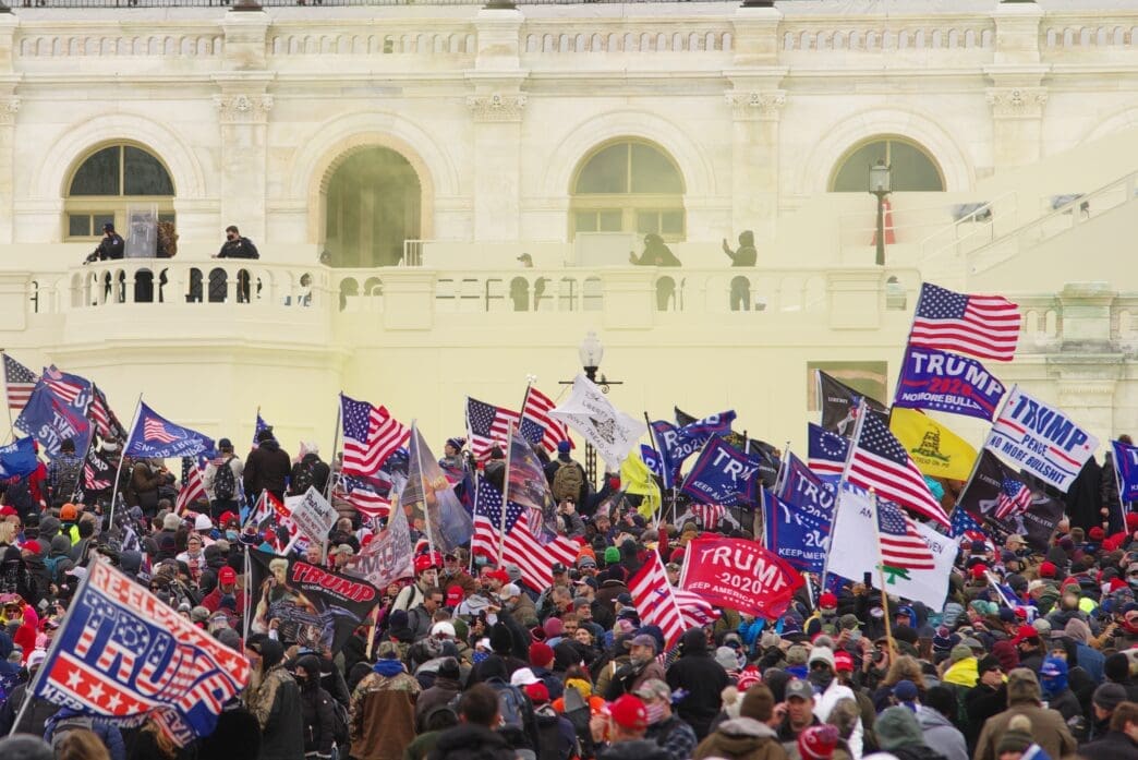 Crowd of Trump supporters with flags gathered near the U.S. Capitol building's exterior with smoke visible.