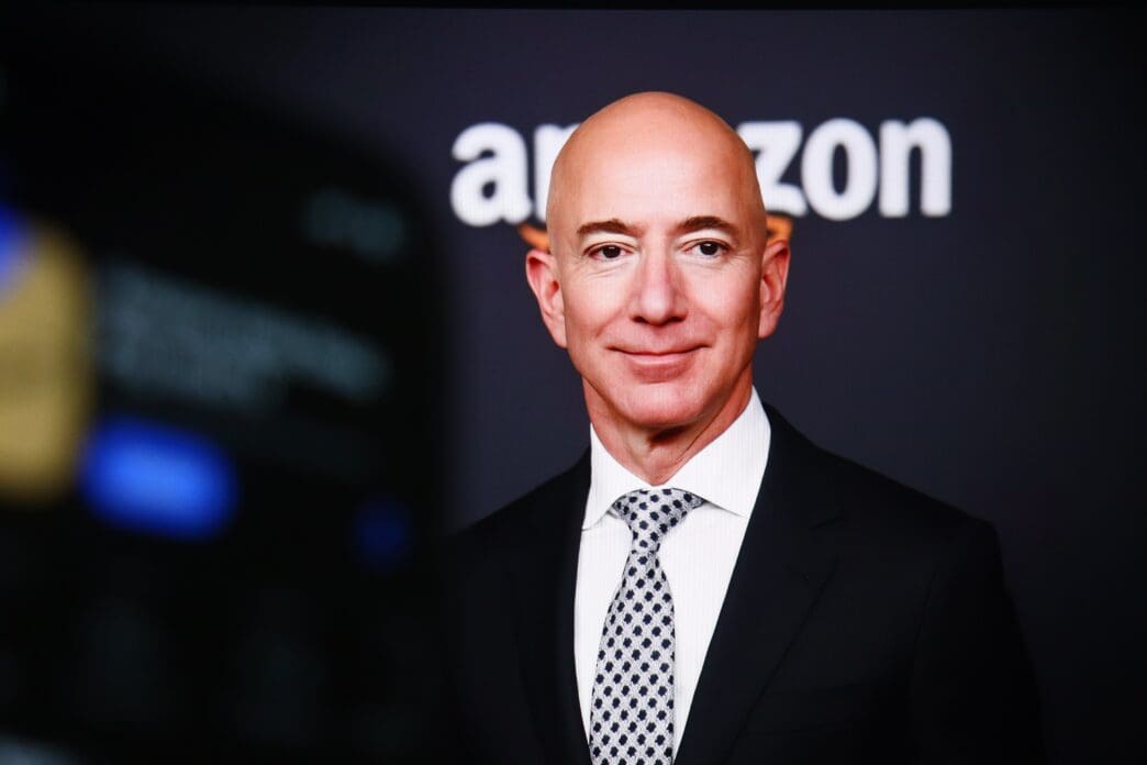 Close-up portrait of Jeff Bezos, bald and smiling, wearing a black suit and a patterned tie, with the Amazon logo visible.
